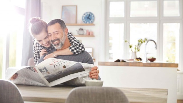 A girl hugs her father, who is sitting at a table reading a newspaper